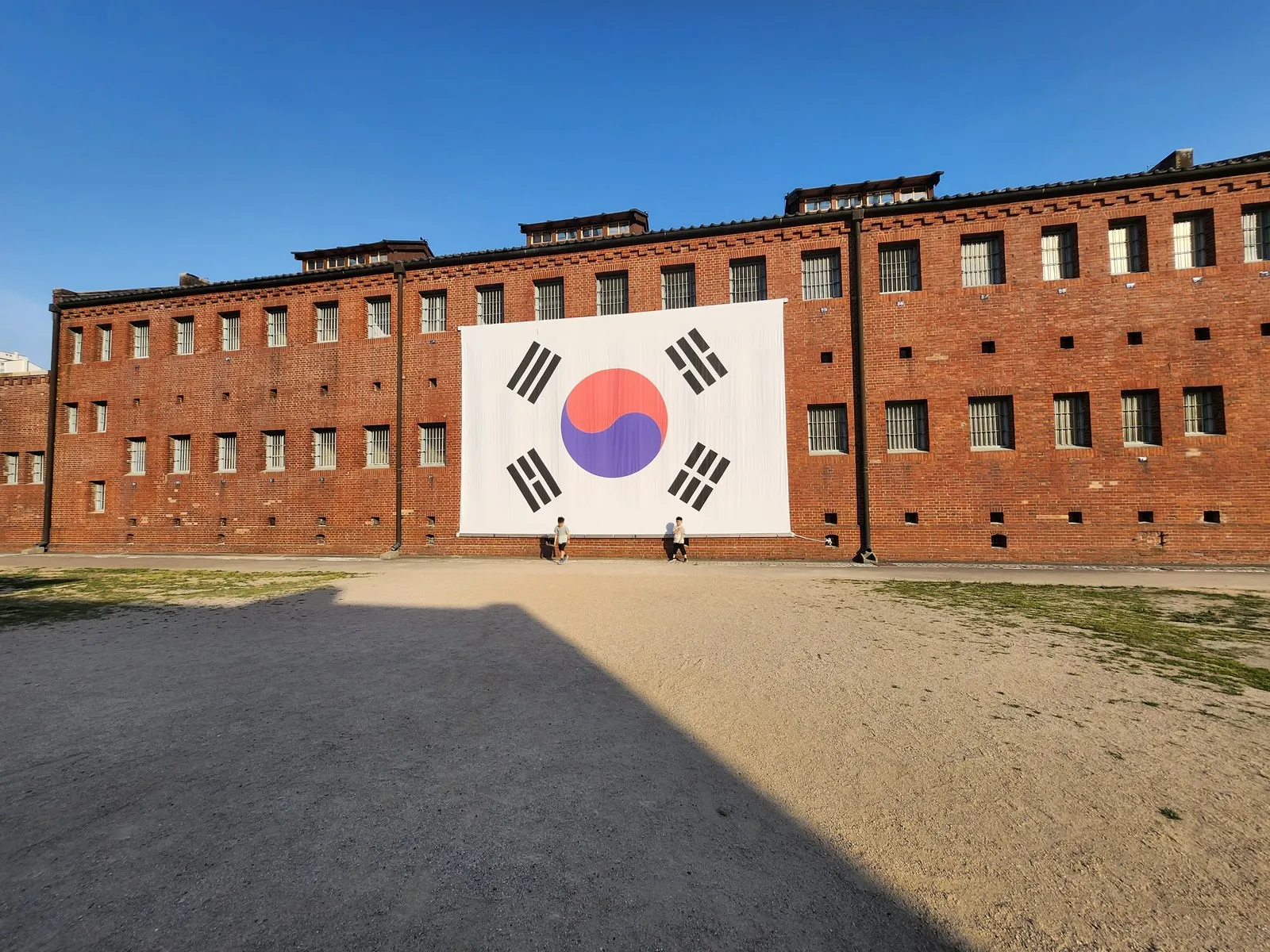 2026년 6월 13일 17:53 — 붉은 벽돌 옥사에 걸린 대형 태극기. 맑은 하늘, 마당을 덮은 긴 오후 그림자